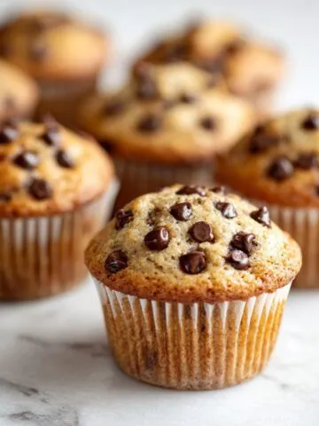 Homemade chocolate chip cupcakes cooling on a white marble counter, imperfectly rustic with chocolate chips on top.