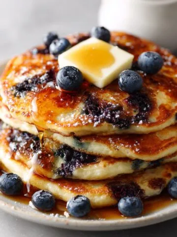 Homemade fluffy blueberry pancakes recipe stacked with butter, maple syrup, and fresh blueberries on a white kitchen counter, overhead shot.