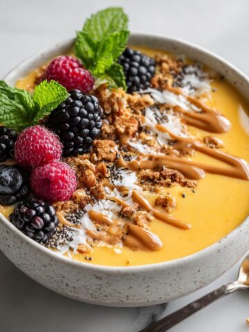 Overhead shot of a homemade mango smoothie bowl recipe topped with granola, berries, chia seeds, and coconut flakes on a white marble kitchen counter.