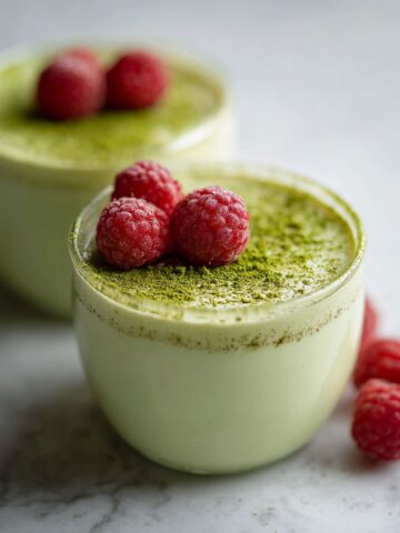 Overhead view of homemade matcha panna cotta recipe in glass ramekins on a white kitchen counter with raspberries and matcha powder, captured in a natural homemade style.