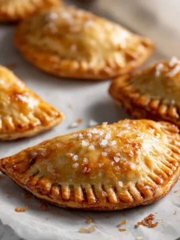 Overhead photo of golden brown mini apple hand pies recipe with rustic crimped edges on parchment paper, sprinkled with sugar, on a white kitchen counter.