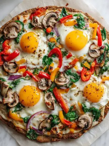 Homemade veggie breakfast pizza recipe with eggs, spinach, bell peppers, mushrooms, and cheese on a white kitchen counter shot from above.