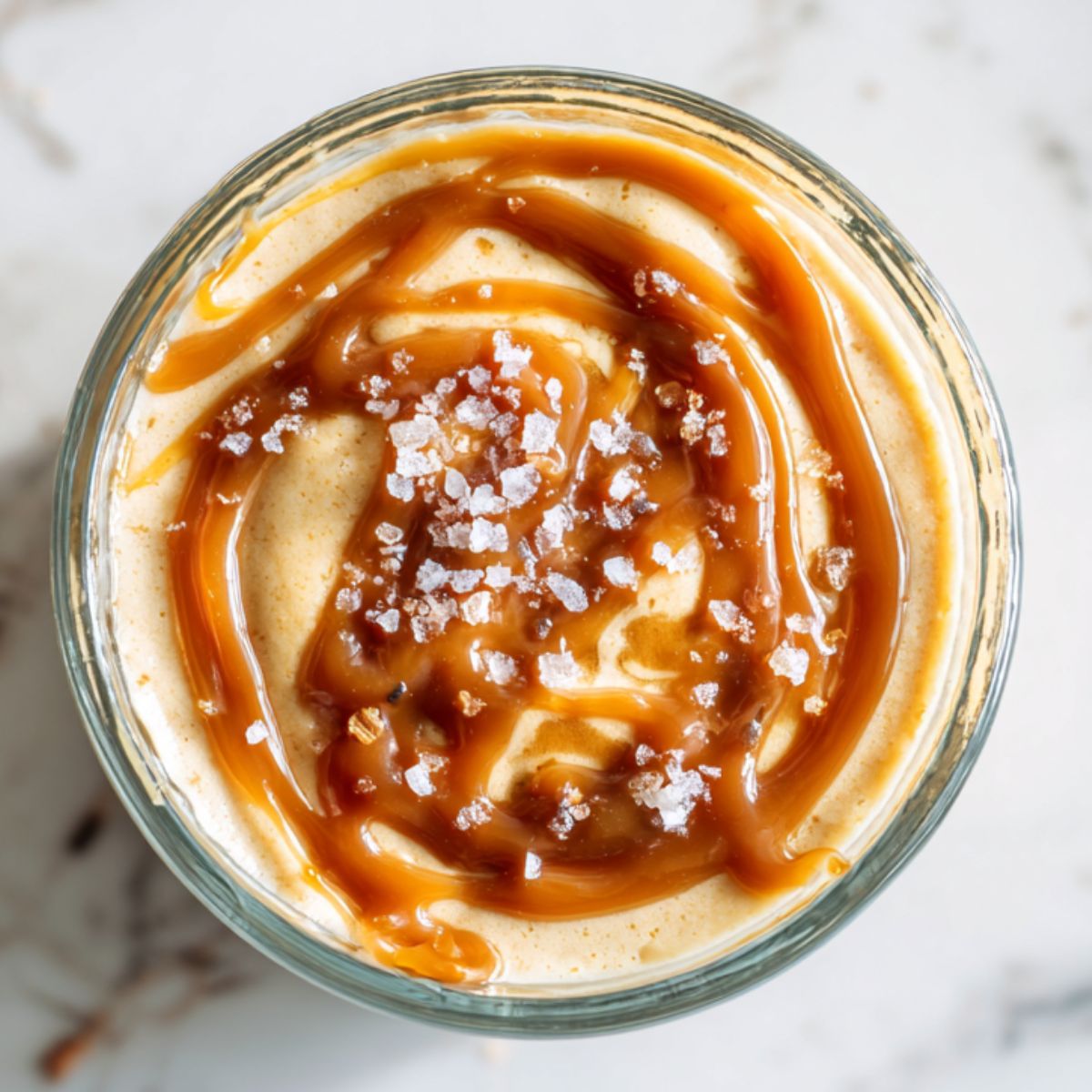 Homemade salted caramel pudding recipe in a glass cup topped with caramel sauce and sea salt.