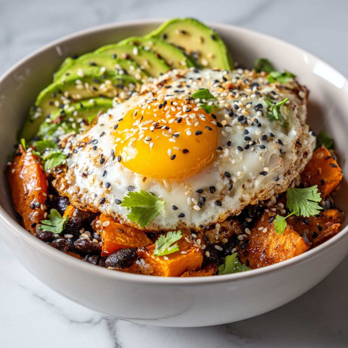 Homemade sweet potato breakfast bowl recipe with avocado, fried egg, black beans, and feta on a white kitchen counter, photographed from above.