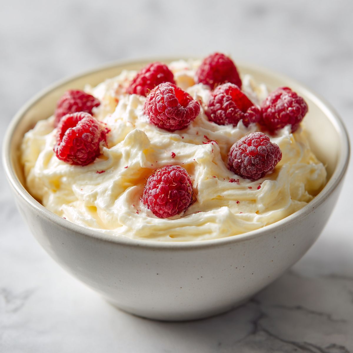 Homemade white chocolate pudding recipe topped with whipped cream and raspberries on a white kitchen counter

