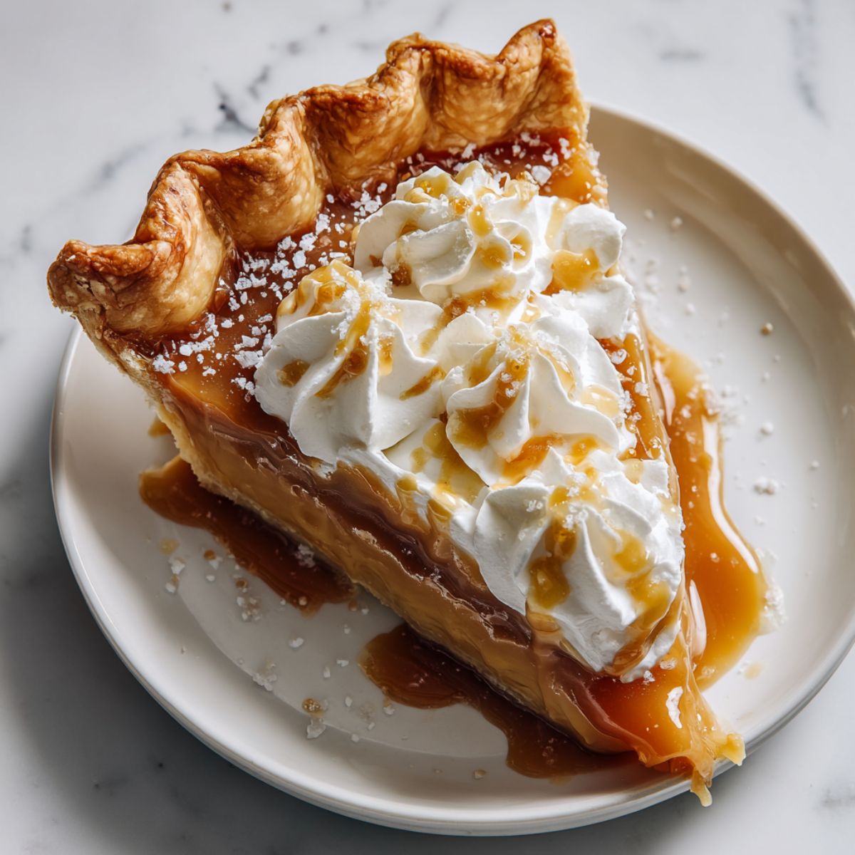 A slice of homemade salted caramel pie recipe topped with whipped cream and sea salt on a white plate.