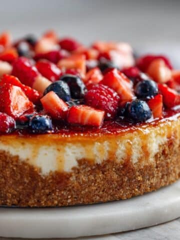 Homemade fruit cheesecake recipe topped with fresh strawberries, blueberries, and raspberries on a white marble counter, photographed overhead with a natural rustic look.