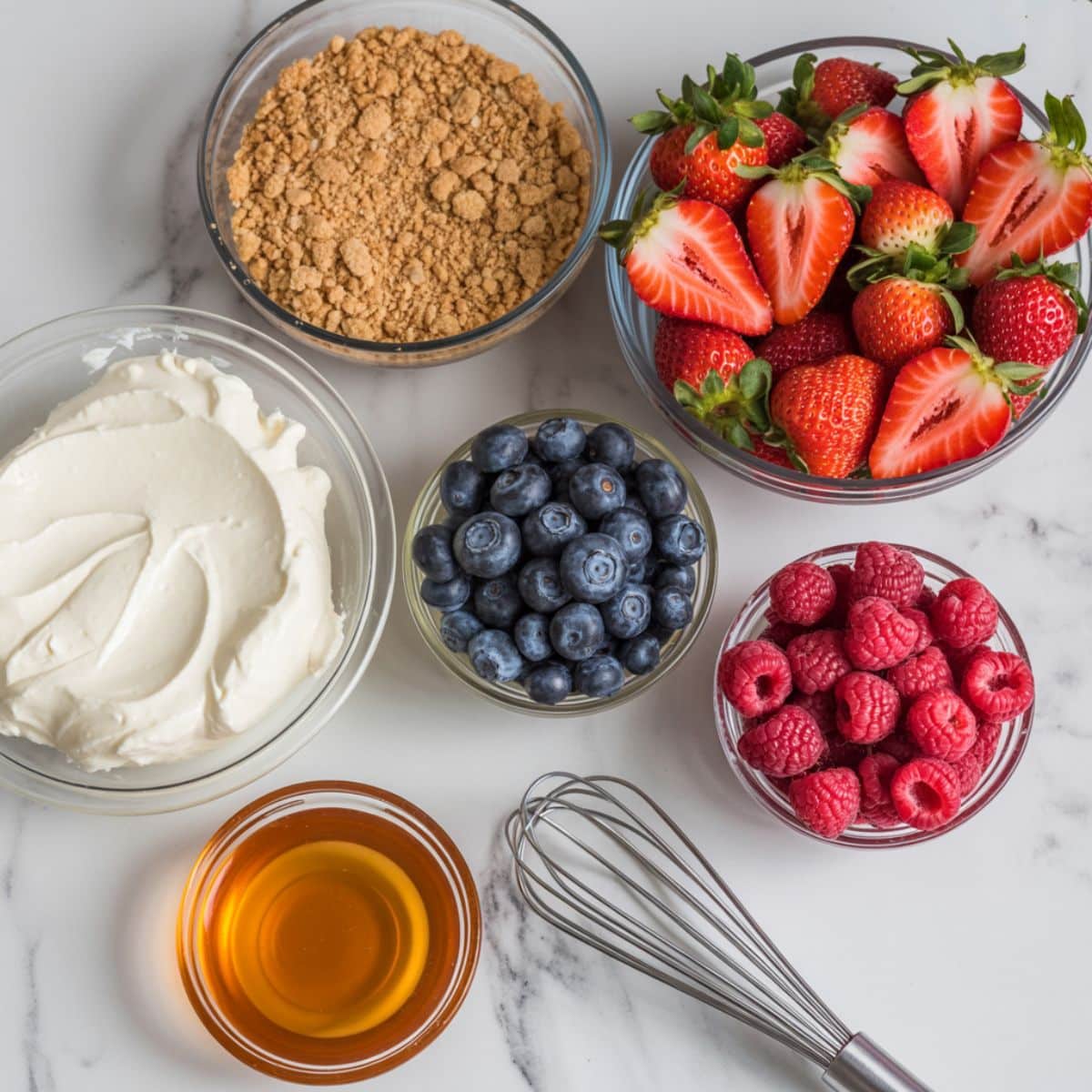 Top-down view of fruit cheesecake ingredients on a white marble kitchen counter including cream cheese, graham cracker crumbs, fresh berries, and honey in a casual homemade setup.