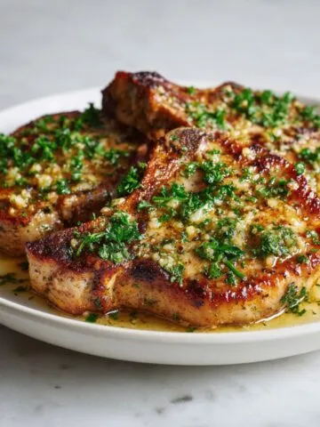 Juicy garlic butter pork chops recipe served on a white plate with sauce and herbs, homemade on a white kitchen counter.