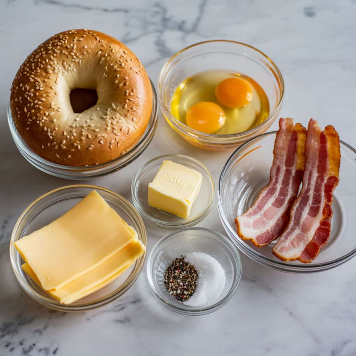 Overhead view of ingredients for a breakfast bagel sandwich including bagel, eggs, cheese, bacon, and butter on a white marble counter.