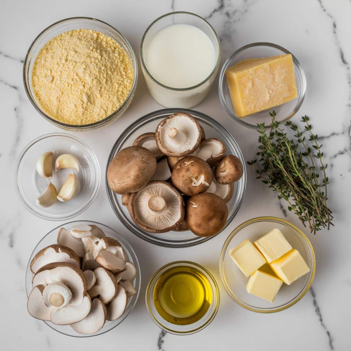 Overhead view of ingredients for creamy polenta with mushrooms, including cornmeal, parmesan, mushrooms, garlic, thyme, butter, and olive oil on a white kitchen counter.