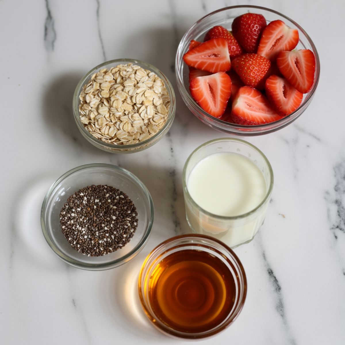 Overhead photo of overnight oats ingredients including rolled oats, chia seeds, strawberries, almond milk, and maple syrup on a white kitchen counter.