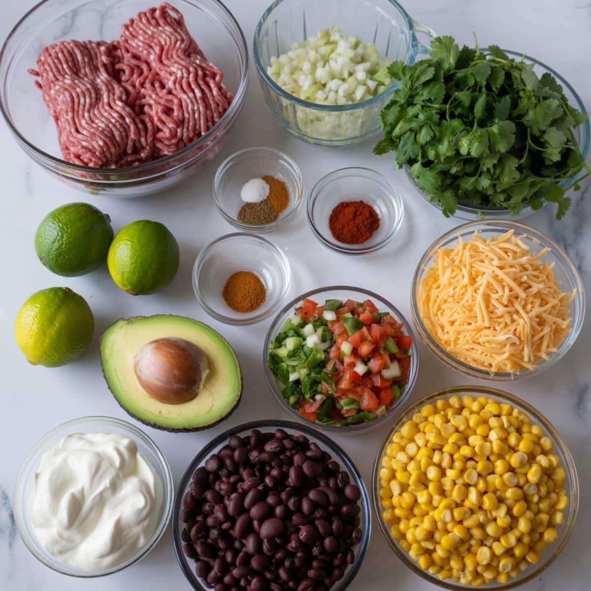 Overhead photo of beef burrito bowl ingredients including ground beef, spices, onion, cilantro, lime, avocado, beans, corn, cheese, pico de gallo, and sour cream on a white counter.