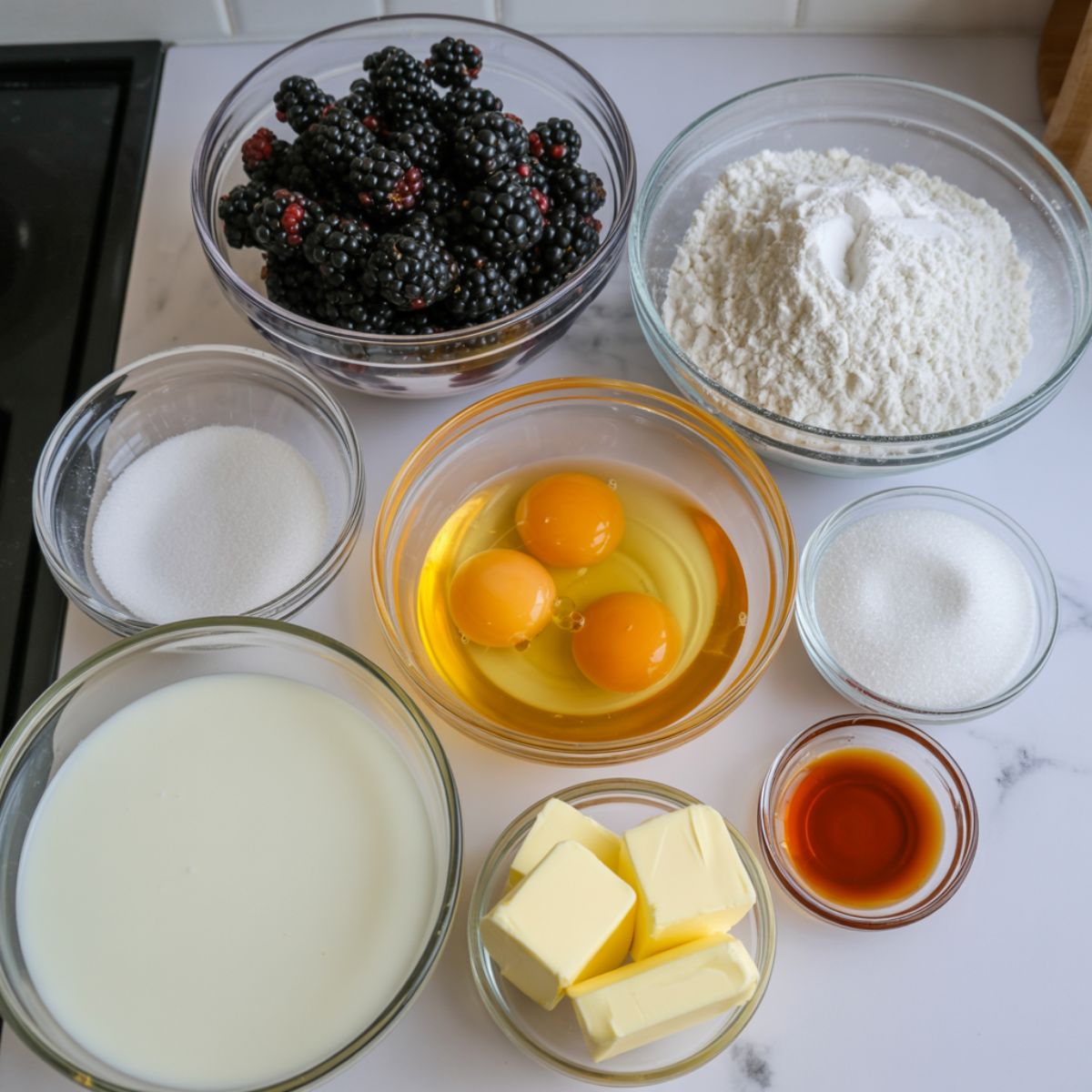 Blackberries, flour, sugar, eggs, butter, and vanilla arranged for baking a cake.