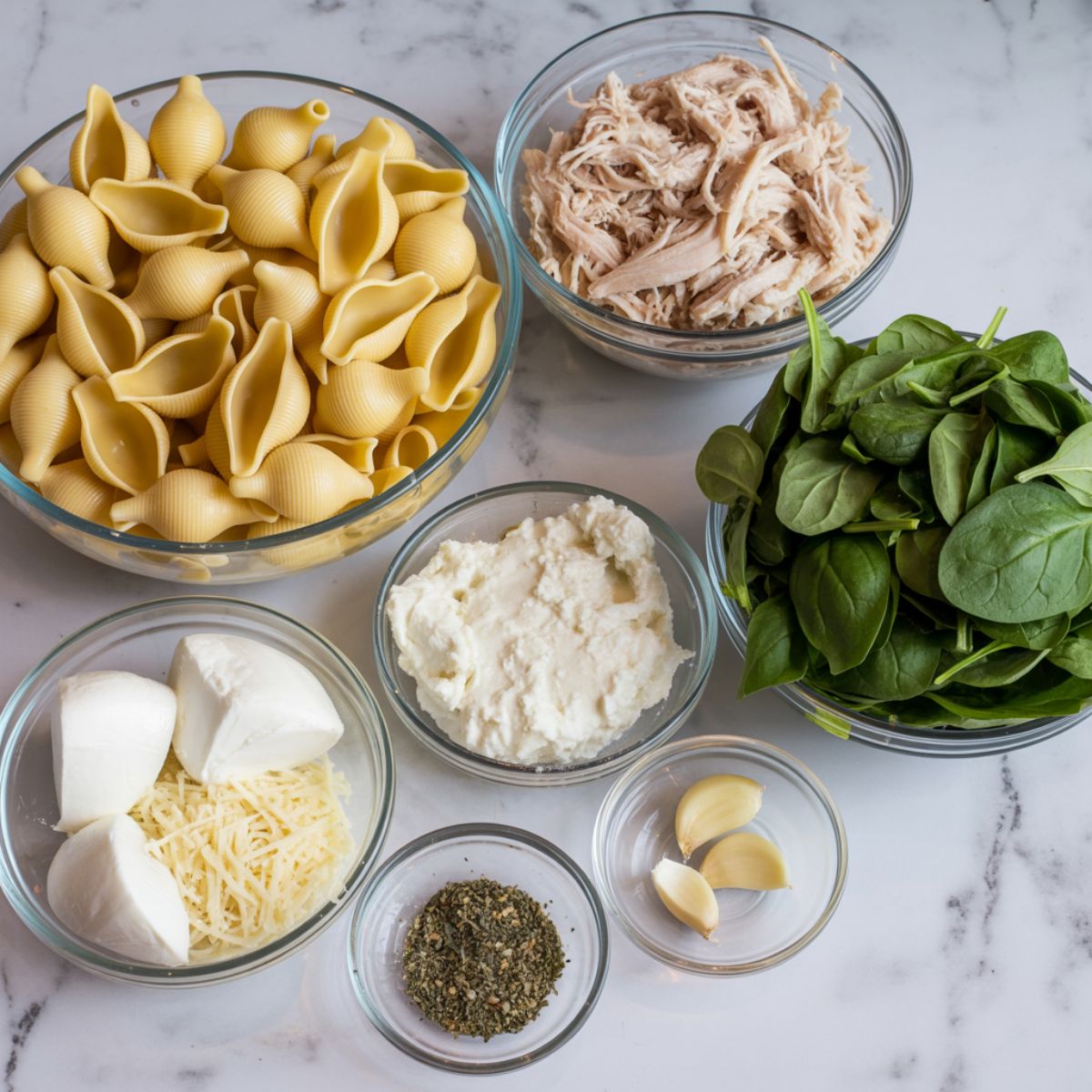 Overhead photo of jumbo pasta shells, shredded chicken, spinach, ricotta, mozzarella, parmesan, and garlic laid out casually on a white marble counter for stuffed shells recipe.