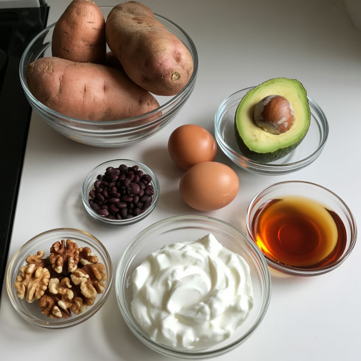 Flat lay of sweet potatoes, eggs, avocado, black beans, yogurt, nuts, and maple syrup on a white kitchen counter, photographed overhead.