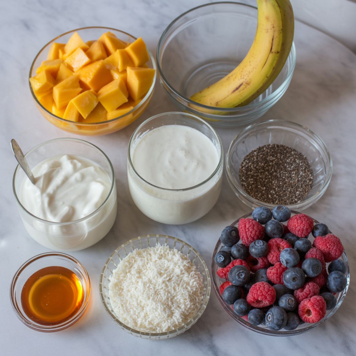 Overhead view of ingredients for a homemade mango smoothie bowl on a white marble counter including mango, banana, coconut milk, yogurt, honey, granola, chia seeds, coconut flakes, and berries.