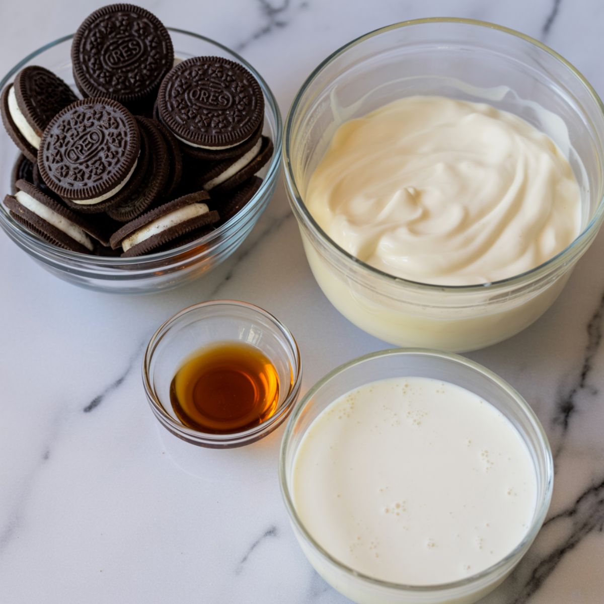 Overhead view of Oreo ice cream ingredients including cookies, condensed milk, vanilla, and cream on a white marble counter.