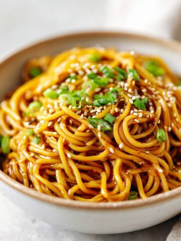 Homemade sesame ginger noodles recipe in a ceramic bowl topped with sesame seeds and green onions on a white kitchen counter, overhead view.