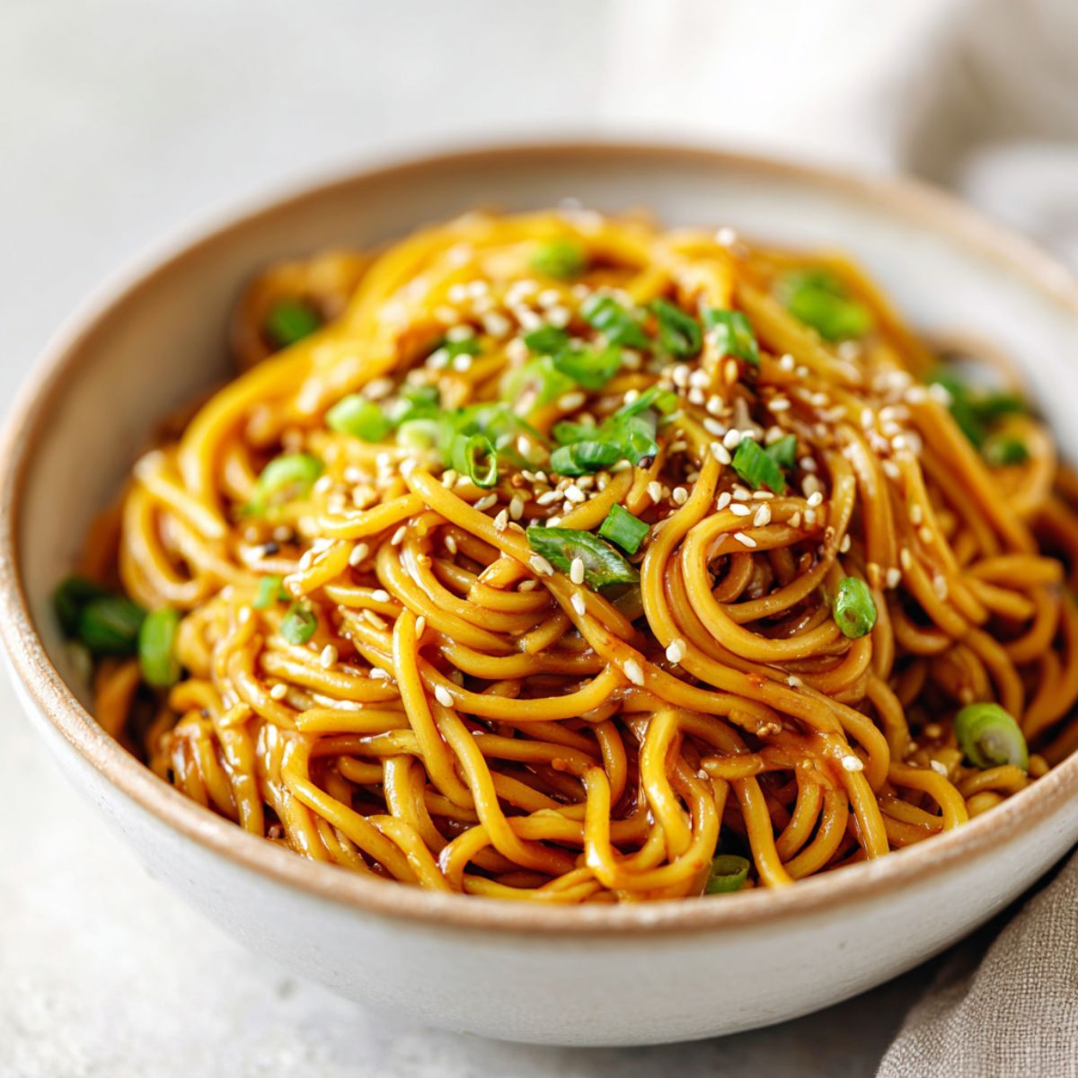 Homemade sesame ginger noodles recipe in a ceramic bowl topped with sesame seeds and green onions on a white kitchen counter, overhead view.