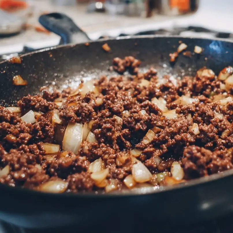 Ground beef cooking in a skillet with onions and garlic for chili on a white stovetop