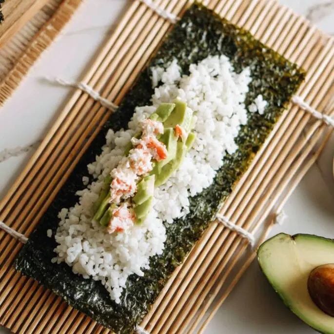 Nori sheet with rice, crab filling, and avocado before rolling.