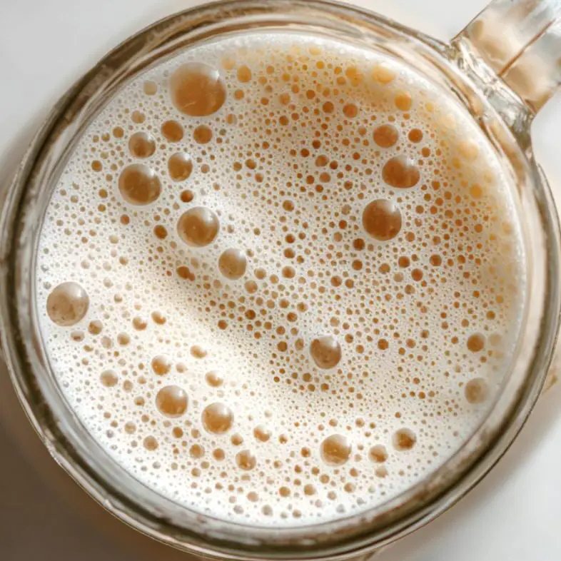Freshly steamed milk with creamy foam in a glass jug on a white kitchen counter.
