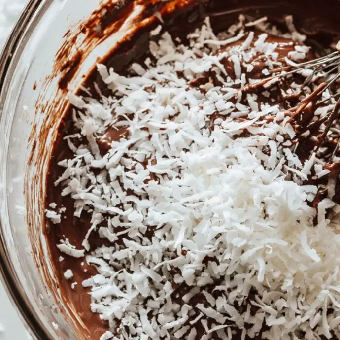 Mixing shredded coconut into brownie batter in a glass bowl
