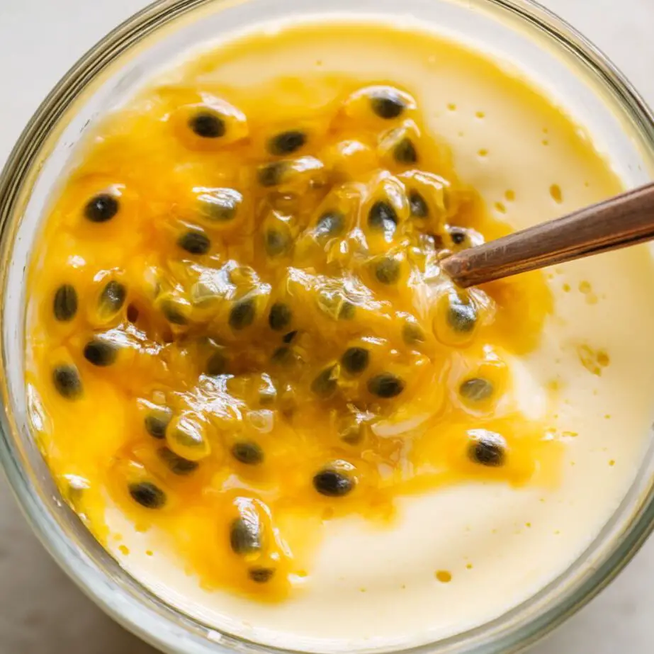 Passion fruit juice being mixed into creamy custard in a glass bowl on a white kitchen counter