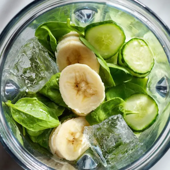 Blender filled with smoothie ingredients ready to blend: banana, spinach, cucumber, and ice

