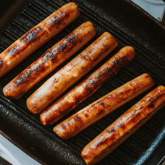 Hot dogs with diagonal cuts browning in a grill pan next to toasted buns