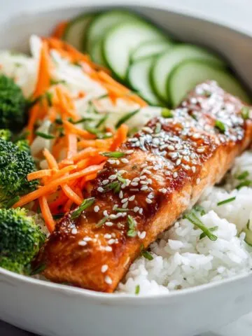 Close-up of a homemade teriyaki salmon rice bowl recipe with glazed salmon, jasmine rice, broccoli, cucumber, and carrots on a white kitchen counter.