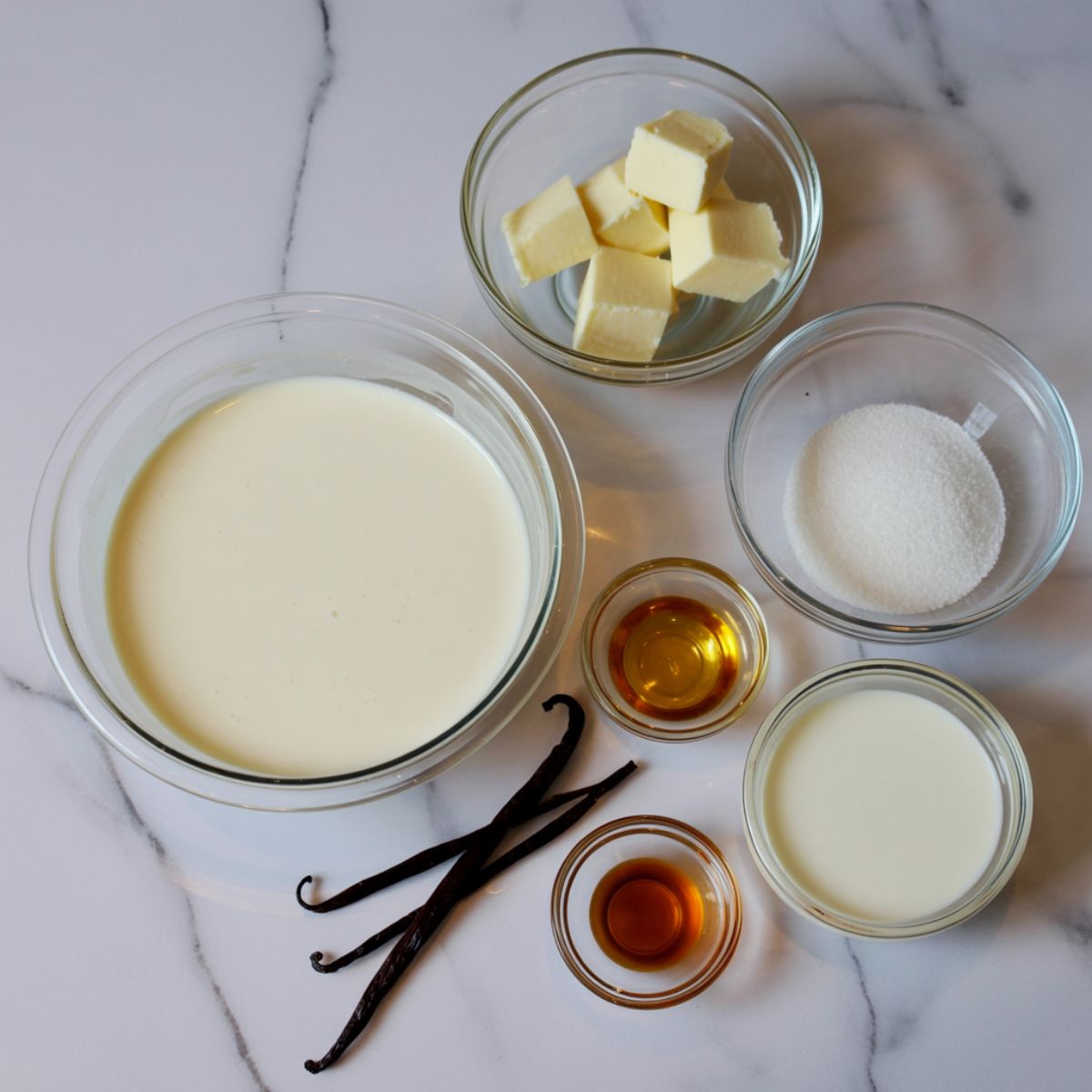Overhead photo of panna cotta ingredients including cream, sugar, vanilla beans, gelatin, and milk on a white marble counter in a homemade setting.