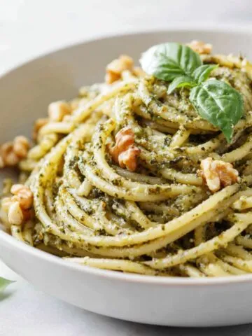 Overhead shot of homemade walnut pesto pasta recipe served in a white bowl on a marble kitchen counter with scattered basil and walnuts