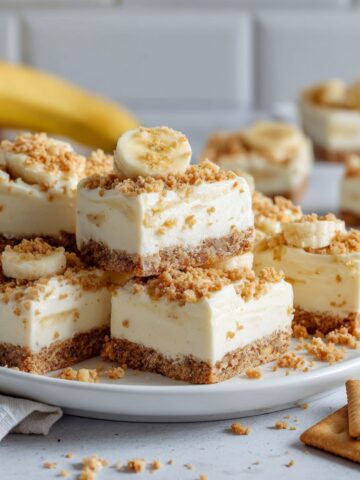 Homemade banana cream bars recipe cut into squares on parchment paper with banana slices and crumbs on a white kitchen counter.