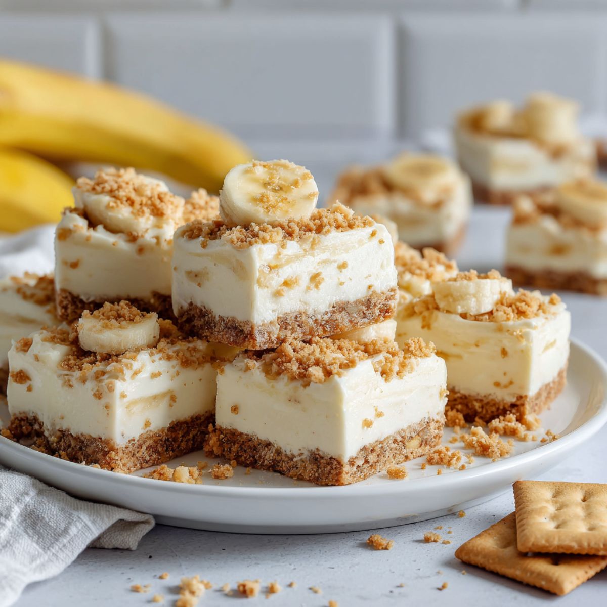 Homemade banana cream bars recipe cut into squares on parchment paper with banana slices and crumbs on a white kitchen counter.