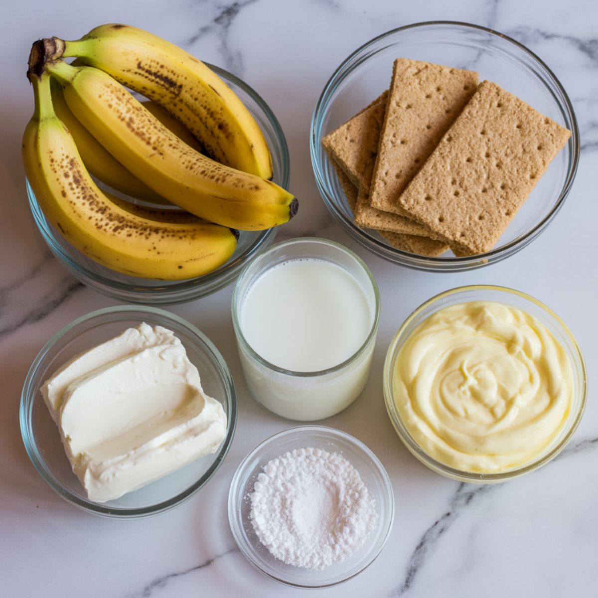 Flat lay of banana cream bars ingredients including bananas, graham cracker crumbs, cream cheese, milk, and pudding mix on a white kitchen counter.
