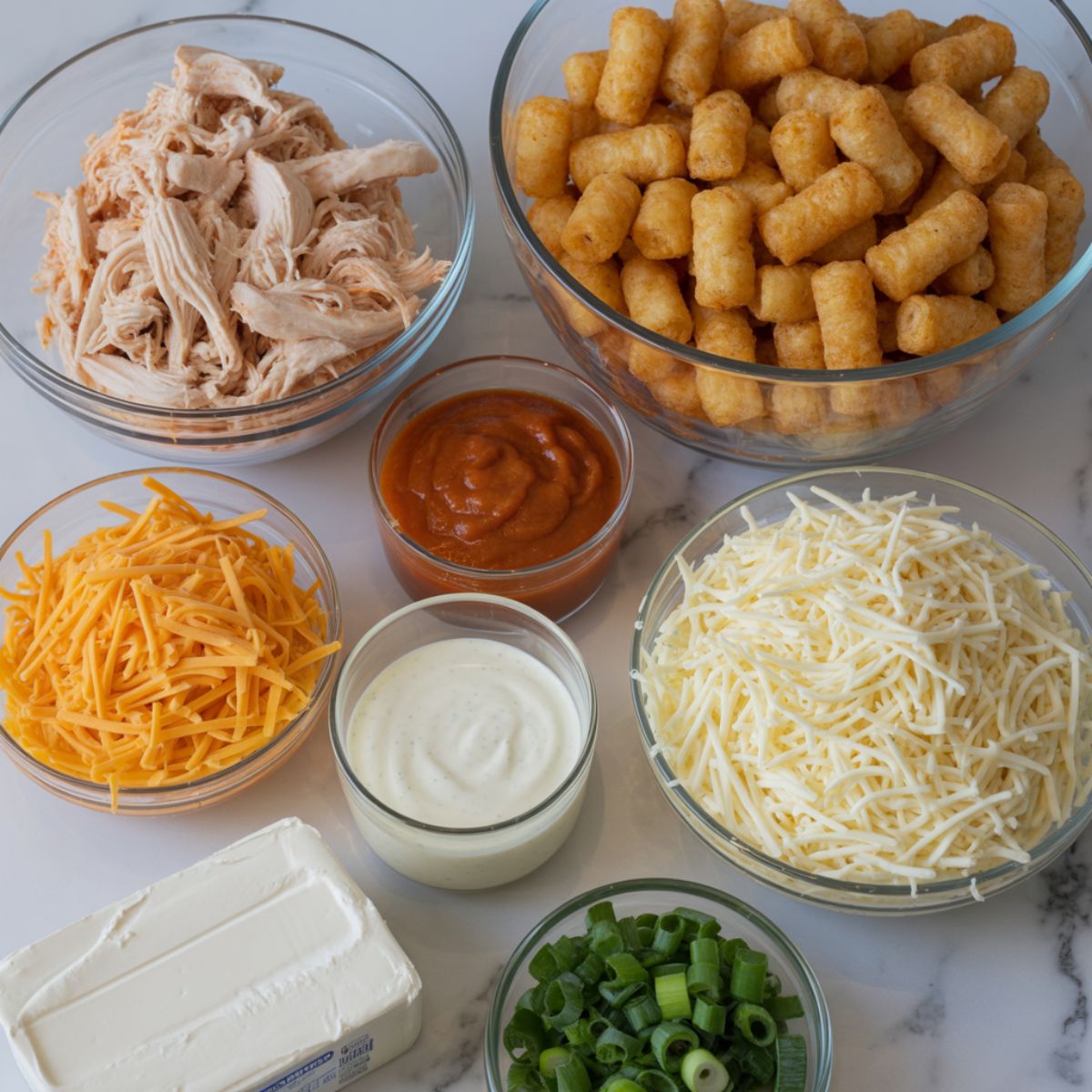 Ingredients for buffalo chicken tater tot casserole laid out on a white kitchen counter, including shredded chicken, frozen tater tots, cheese, buffalo sauce, ranch, and green onions, photographed from above.