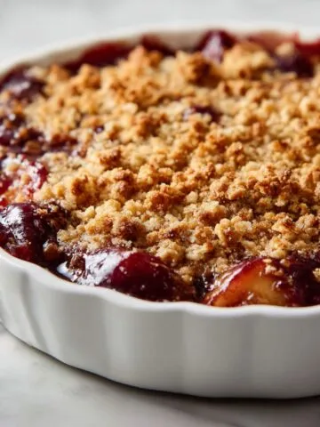 Homemade plum crisp recipe in a white ceramic baking dish with golden oat topping and bubbling plum filling, photographed overhead on a white kitchen counter.