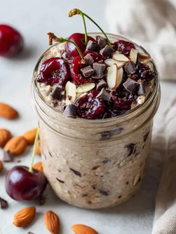 Homemade jar of chocolate cherry overnight oats recipe topped with cherries, chocolate chips, and almonds on a white kitchen counter, overhead view.