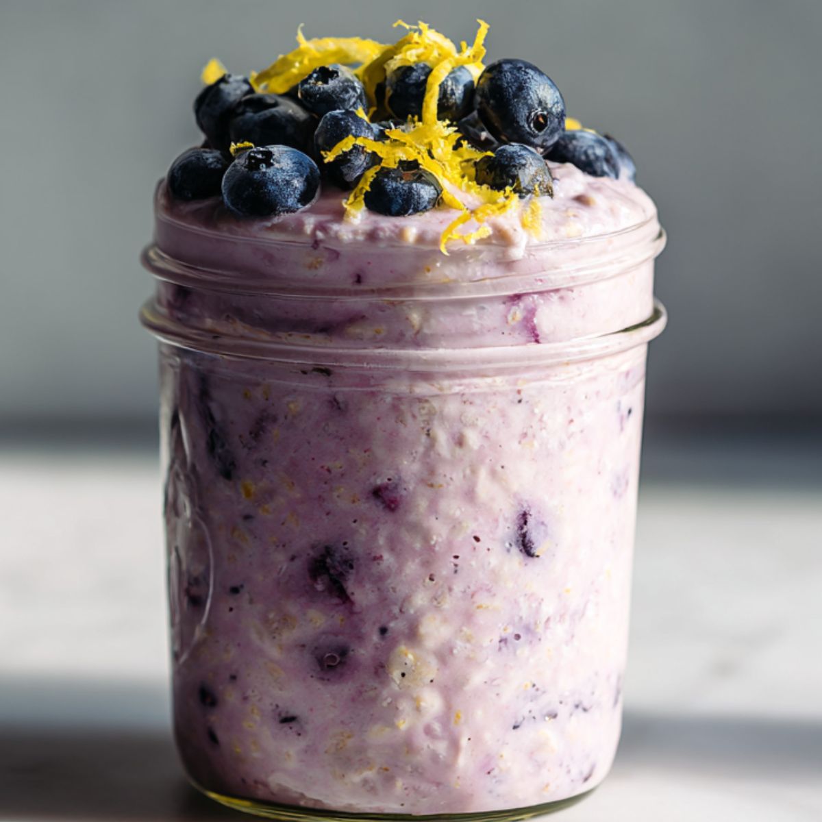 Overhead shot of a homemade jar of blueberry lemon overnight oats recipe topped with fresh blueberries and lemon zest on a white kitchen counter.