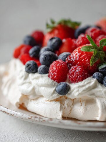Homemade mixed berry pavlova recipe topped with fresh cream and berries on a white kitchen counter, photographed from above.