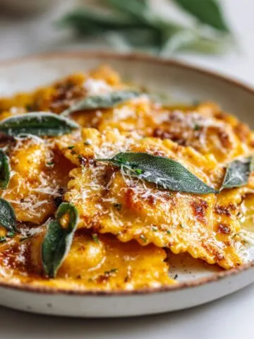Overhead shot of butternut squash ravioli recipe ingredients including squash cubes, sage, parmesan, ricotta, eggs, and flour on a white kitchen counter.