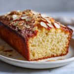 Overhead view of a homemade French butter cake recipe loaf with golden crust and almonds on a white kitchen counter.
