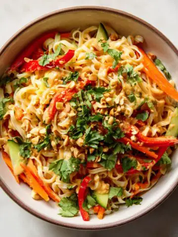 Overhead view of a homemade peanut noodle bowl recipe with creamy peanut sauce, fresh vegetables, and peanuts on a white kitchen counter.