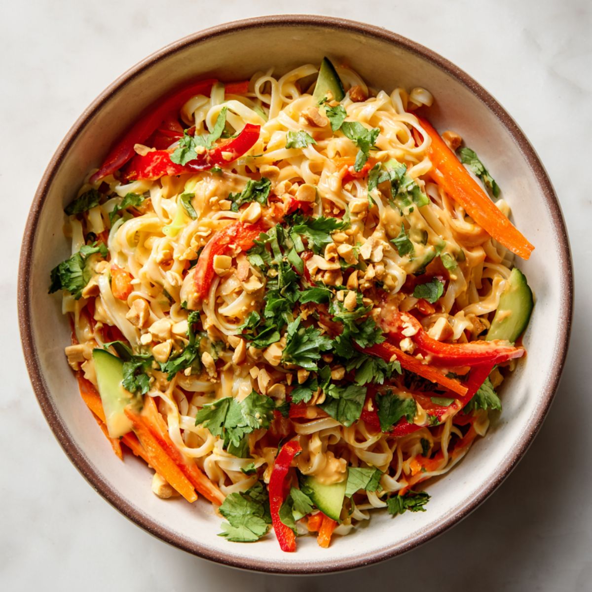 Overhead view of a homemade peanut noodle bowl recipe with creamy peanut sauce, fresh vegetables, and peanuts on a white kitchen counter.