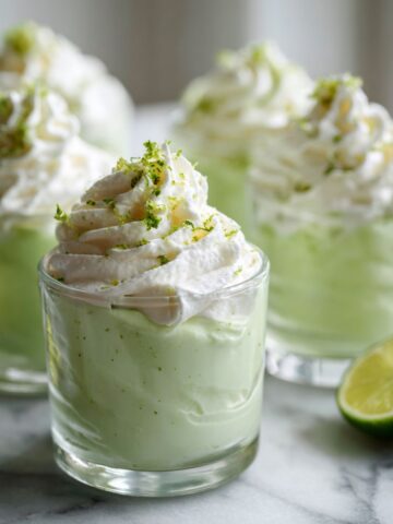 Homemade key lime mousse cups recipe in clear glasses topped with whipped cream and lime zest, photographed overhead on a white kitchen counter.