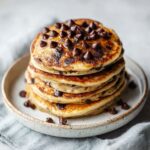 Stack of homemade chocolate chip protein pancakes recipe on a white kitchen counter with melted chocolate chips on top, photographed from above in a natural, homemade style.