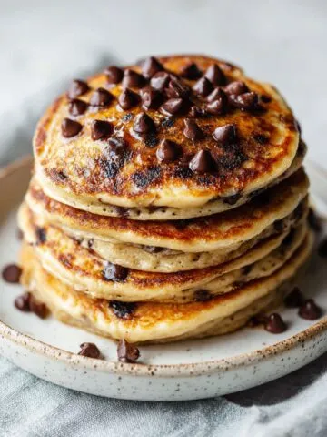 Stack of homemade chocolate chip protein pancakes recipe on a white kitchen counter with melted chocolate chips on top, photographed from above in a natural, homemade style.