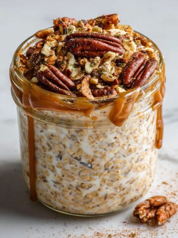 Homemade pecan pie overnight oats recipe in a glass jar topped with pecans and cinnamon on a white marble counter.